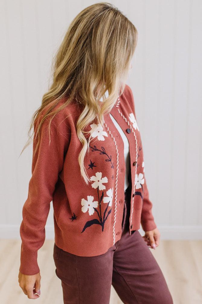 A woman wearing a terracotta-colored knit cardigan with white floral embroidery and dark leaf details, paired with dark brown pants, shown from a side-back angle against a light wood-paneled wall.