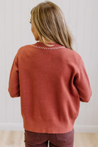 A woman viewed from behind wearing a rust-colored knit sweater with a white zigzag border on the collar, paired with dark brown pants, set against a white paneled wall.