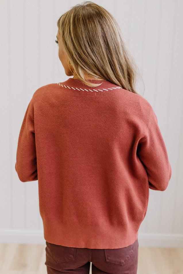 A woman viewed from behind wearing a rust-colored knit sweater with a white zigzag border on the collar, paired with dark brown pants, set against a white paneled wall.