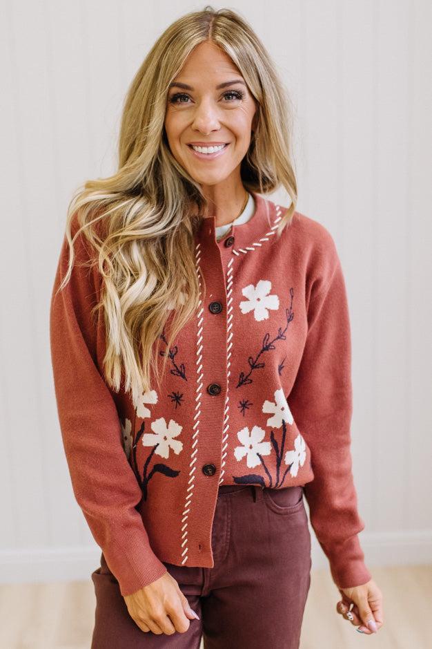 A smiling blonde woman wearing a rust-colored knit cardigan with white floral embroidery and dark buttons, paired with dark brown pants, standing against a light paneled wall.