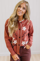 A woman modeling a rust-colored cardigan with white floral embroidery and a white top, paired with dark brown wide-leg pants and white pointed-toe heels, standing in a bright room with light wood floors and white paneled walls.