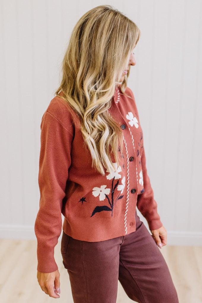 A woman wearing a rust-colored knit cardigan sweater with white floral embroidery and dark buttons, paired with dark brown pants, standing against a white paneled wall.