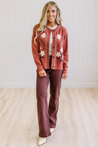A woman modeling a rust-colored cardigan with white floral embroidery and a white top, paired with dark brown wide-leg pants and white pointed-toe heels, standing in a bright room with light wood floors and white paneled walls.