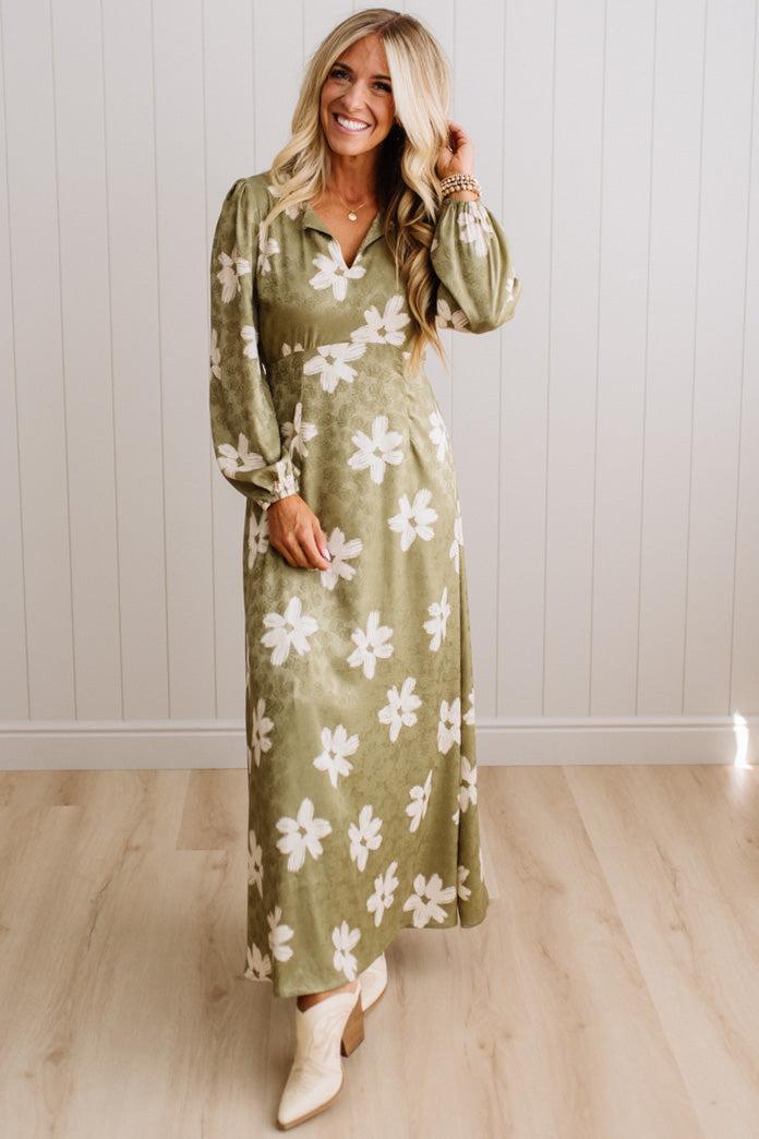 A woman modeling a long-sleeve olive green maxi dress with a white floral pattern, standing in front of a white paneled wall and light wood floor.
