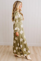 A woman modeling a long-sleeve olive green maxi dress with a white floral pattern, standing in front of a white paneled wall and light wood floor.