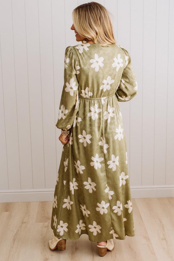 A woman wearing a long-sleeved olive green maxi dress with a white floral pattern, shown from the back against a white paneled wall and light wood floor.