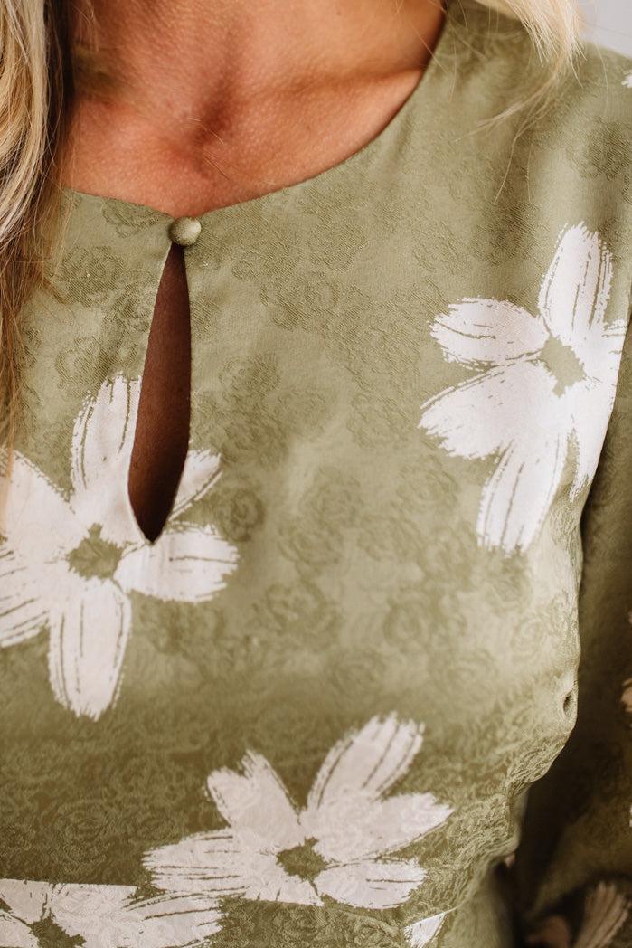 Close-up of a person wearing a sage green dress with a white floral pattern and a keyhole neckline with a brown tie detail.