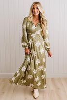A woman modeling a long-sleeve olive green maxi dress with a white floral pattern, standing in front of a white paneled wall and light wood floor.