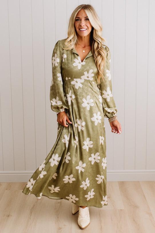 A woman modeling a long-sleeve olive green maxi dress with a white floral pattern, standing in front of a white paneled wall and light wood floor.