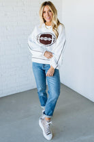 Woman wearing a white sweater with a graphic design, blue jeans, and white sneakers against a white brick wall.