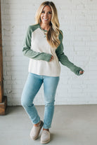 Woman wearing a green and white long-sleeve shirt with blue jeans against a white brick wall.