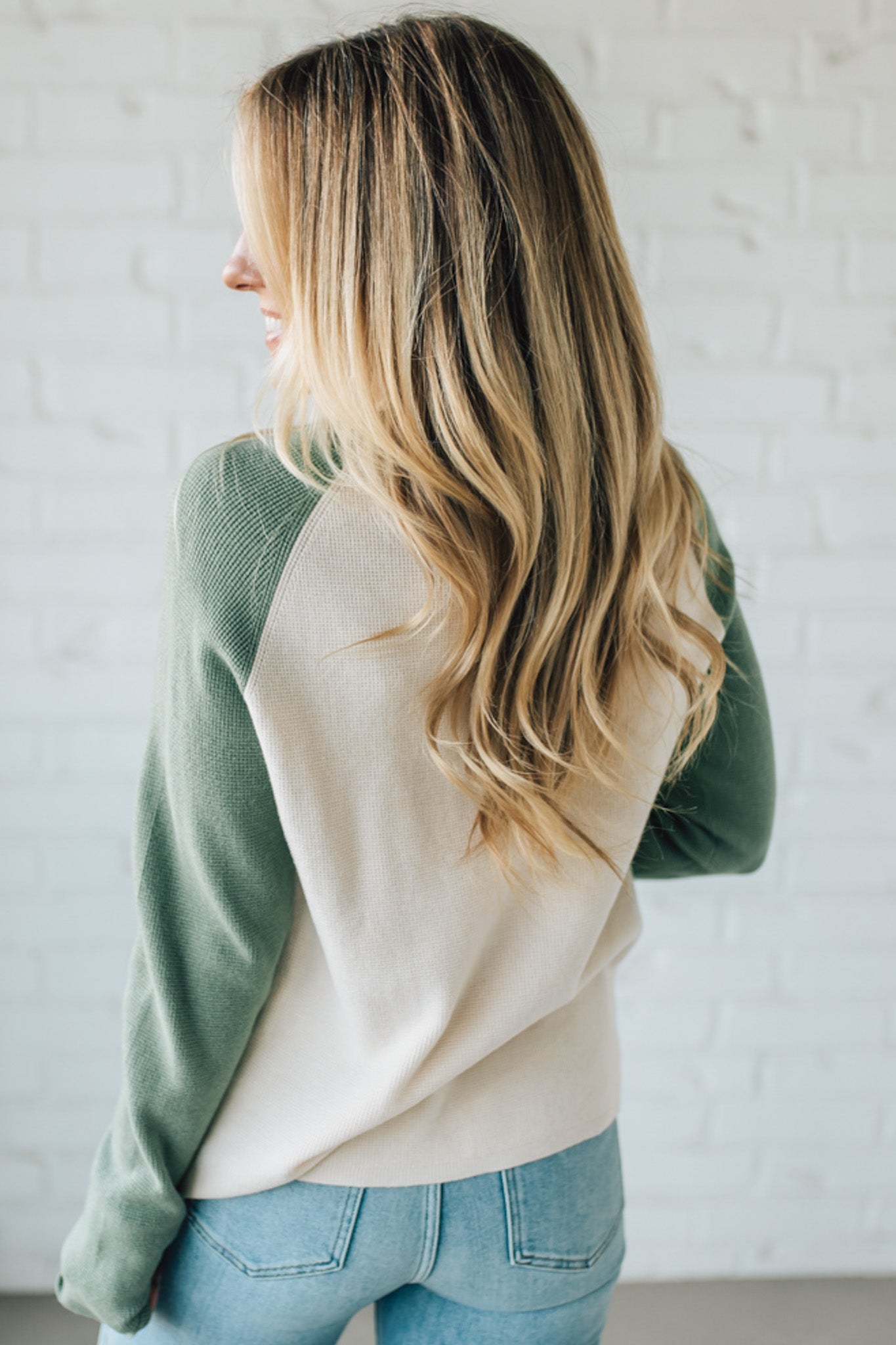 Woman wearing a green and beige sweater with blue jeans against a white wall.
