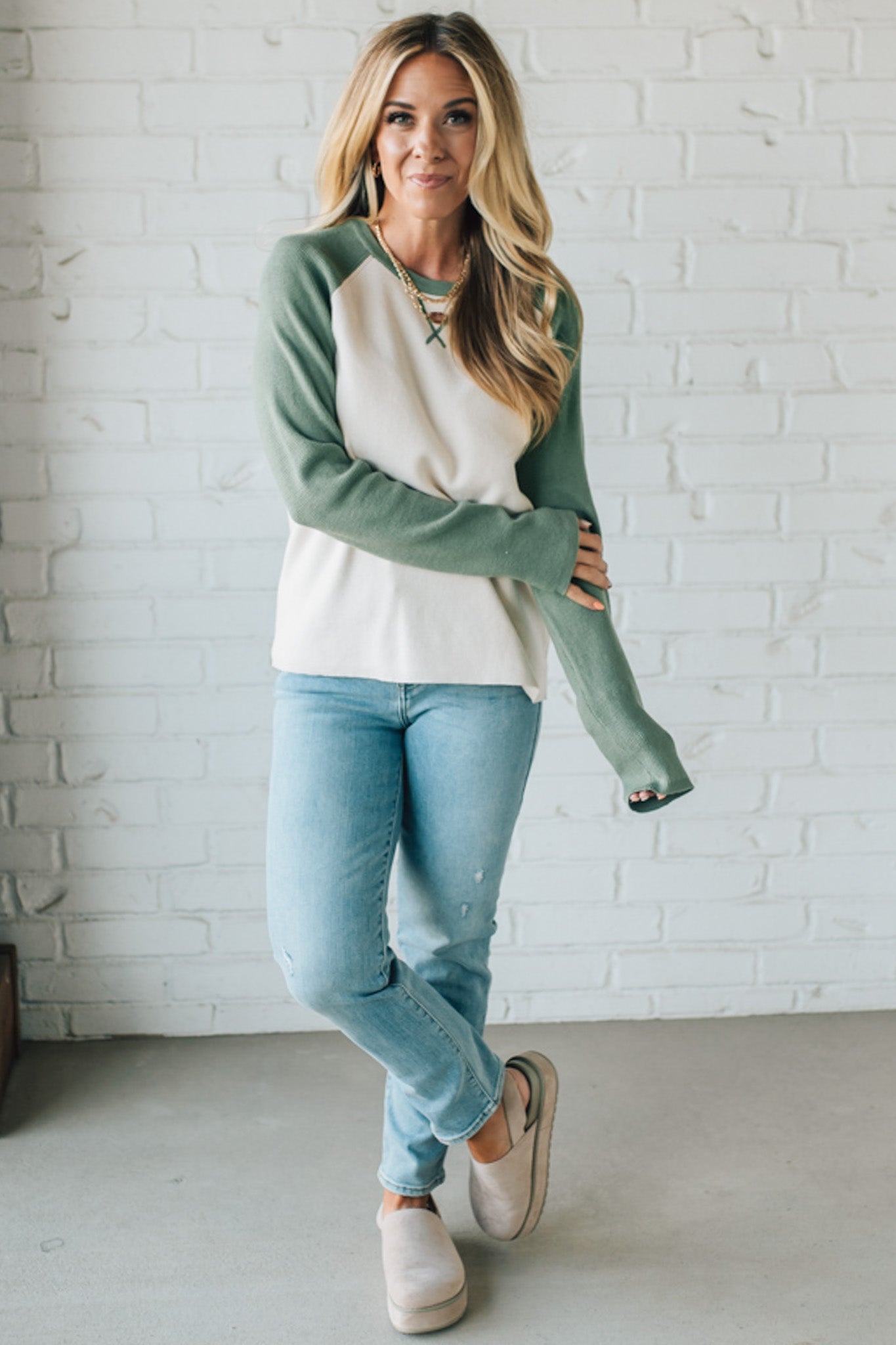 Woman wearing a green and white long-sleeve shirt with light blue jeans against a white brick wall.
