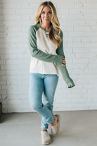 Woman wearing a green and white long-sleeve shirt with light blue jeans against a white brick wall.