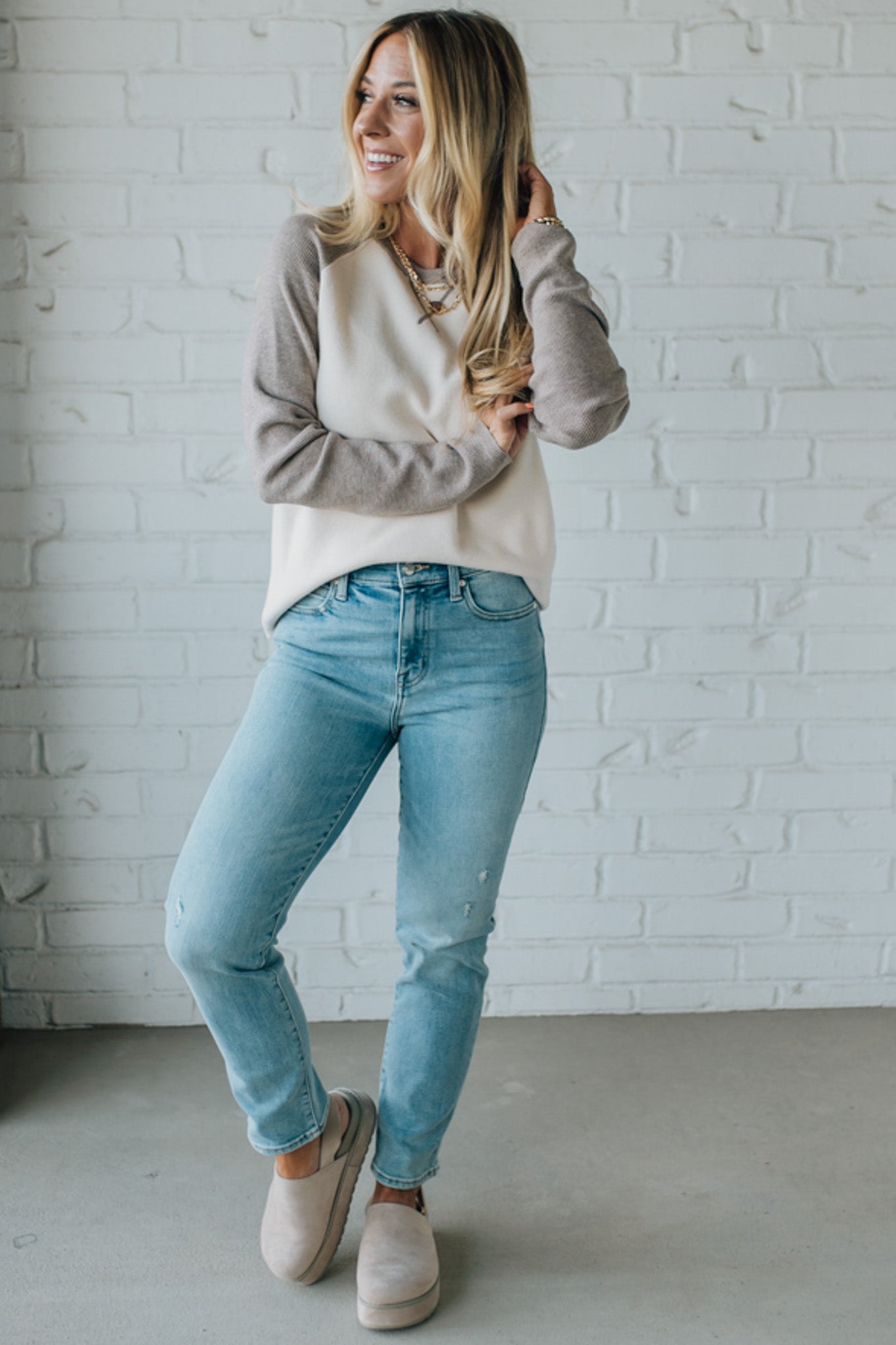 Woman wearing a beige sweater and light blue jeans against a white brick wall.