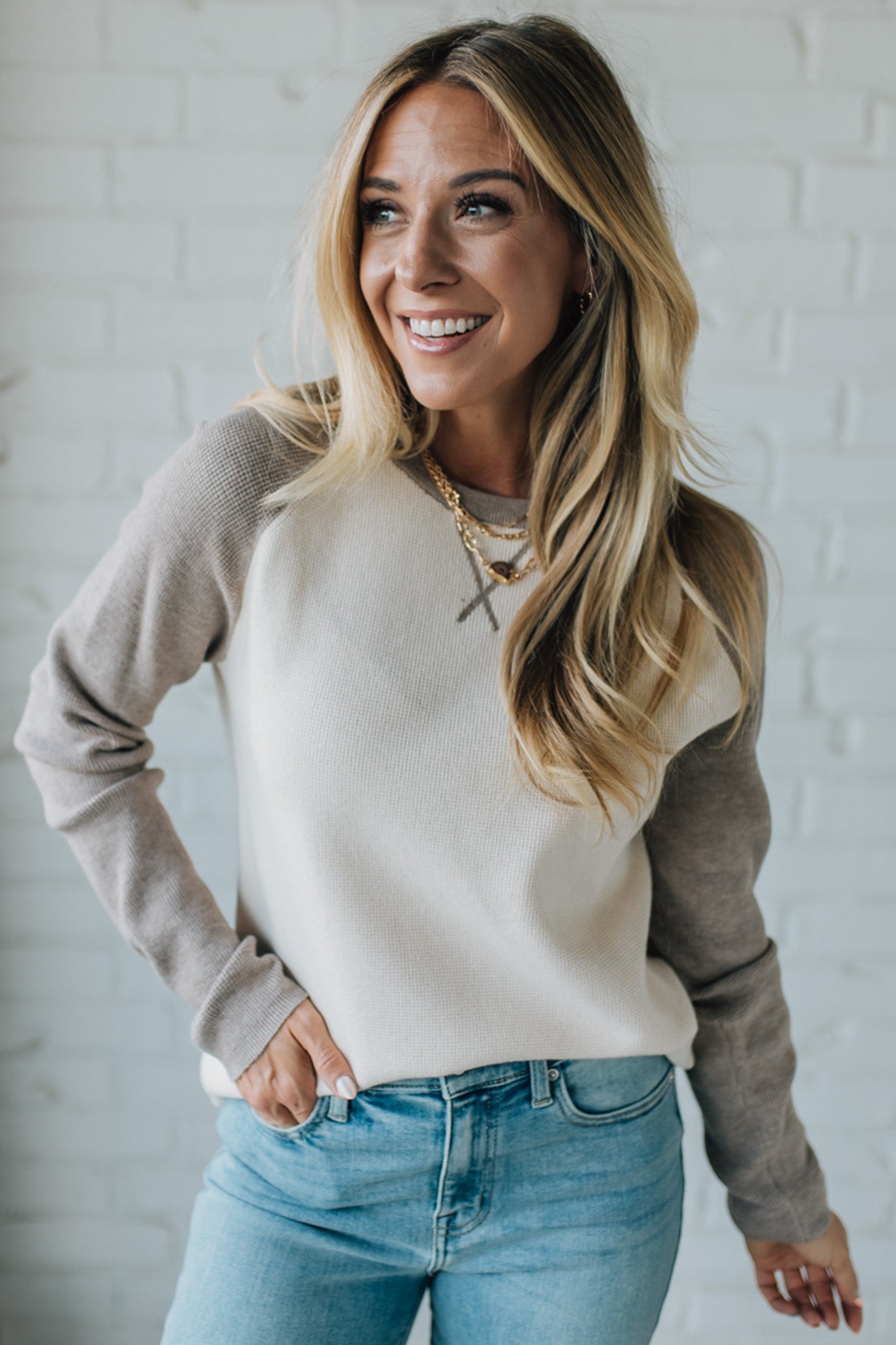 Woman wearing a beige and brown sweater with blue jeans against a white brick wall.