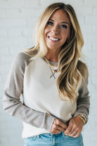 Woman wearing a beige sweater and light blue jeans against a white brick wall.