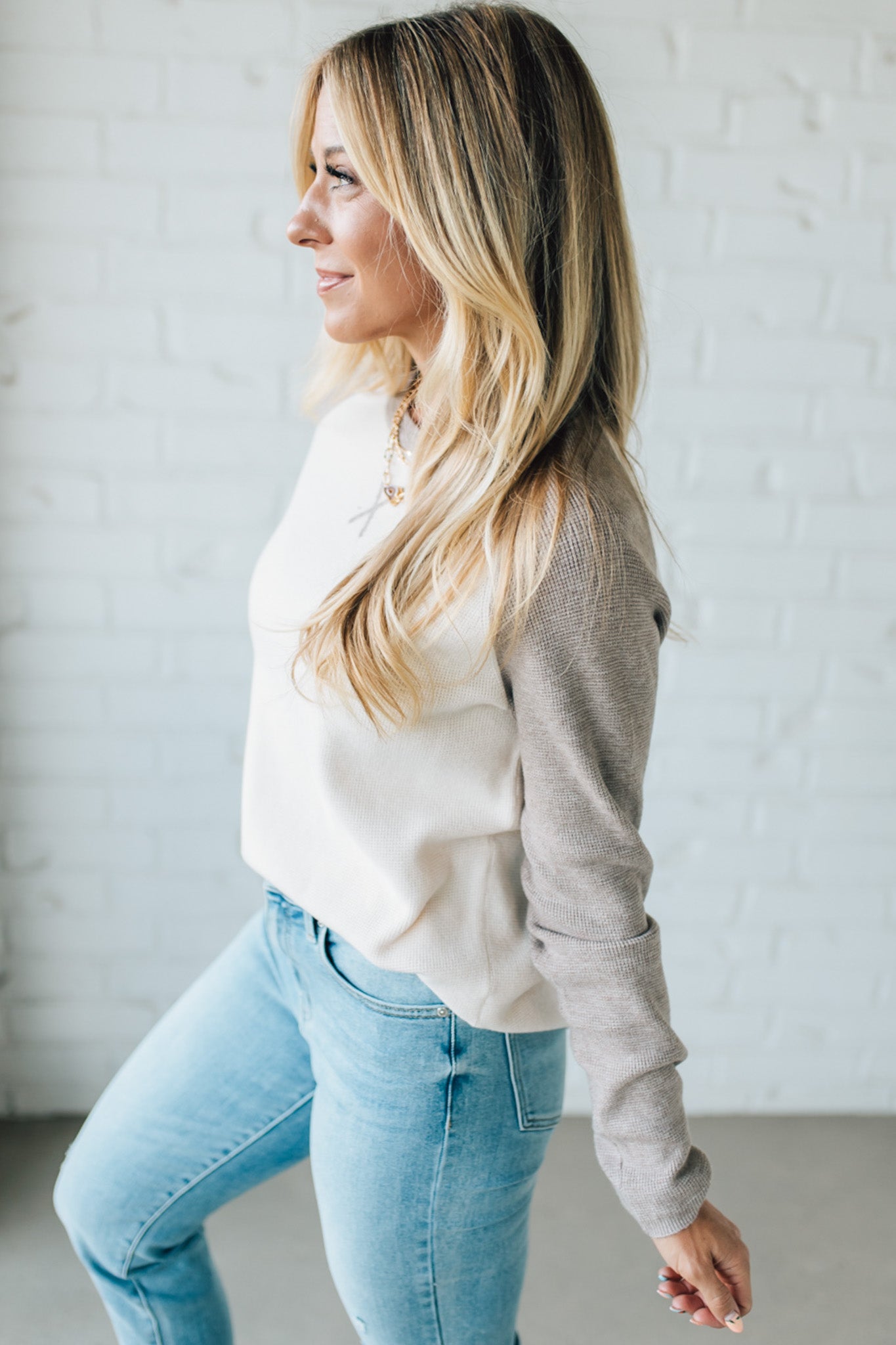 Woman wearing a white top and light blue jeans against a white brick wall.