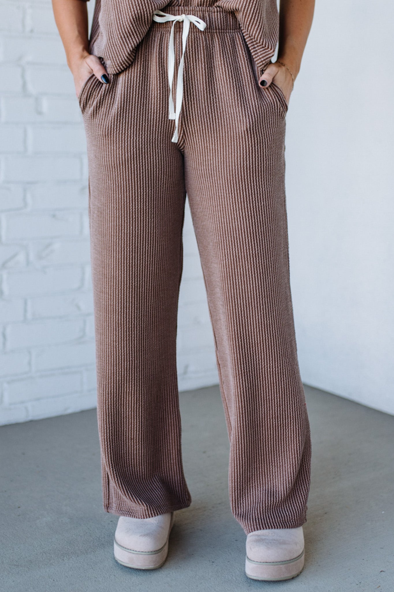 Brown ribbed pants with a white drawstring against a white brick wall.