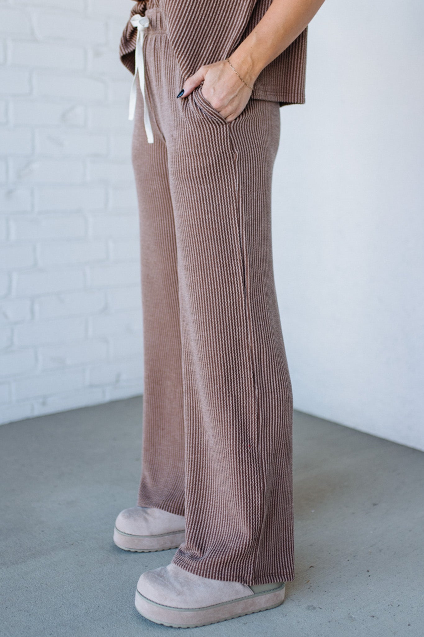 Person wearing brown ribbed pants and white sneakers against a white brick wall.