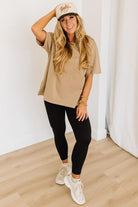 Woman wearing a beige t-shirt, black leggings, and white sneakers with a white cap indoors.