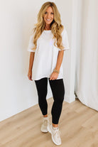 Woman wearing a white t-shirt, black leggings, and white sneakers in a minimalistic setting.