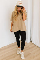 Woman wearing a beige t-shirt, black leggings, and white sneakers with a beige cap indoors.