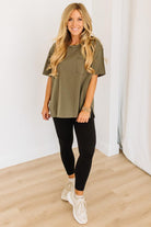 Woman wearing an olive green top and black leggings standing in a room with white walls and wooden floor.