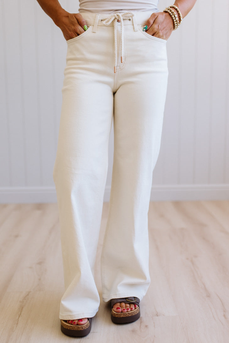 Person wearing white flared jeans and sandals on a light wooden floor.