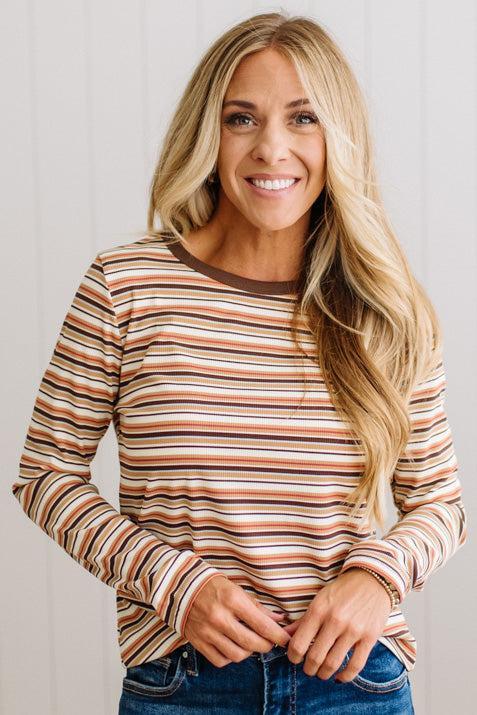A smiling blonde woman wearing a long-sleeve crewneck shirt with horizontal stripes in rust orange, brown, cream, and white, paired with blue jeans, set against a light gray paneled wall.