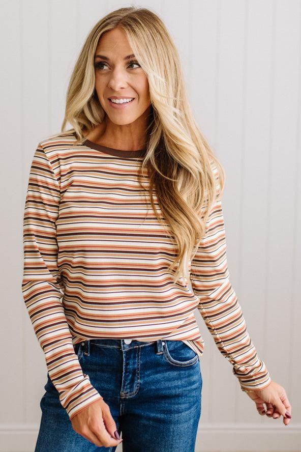 A woman with long blonde hair wearing a long-sleeve crewneck top with horizontal stripes in brown, orange, and white, paired with blue jeans, standing against a white paneled wall.