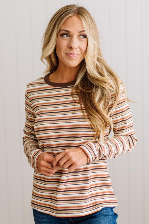A woman with long blonde hair wearing a long-sleeve crewneck shirt with horizontal stripes in brown, orange, beige, and white, paired with blue jeans, standing against a white paneled wall.