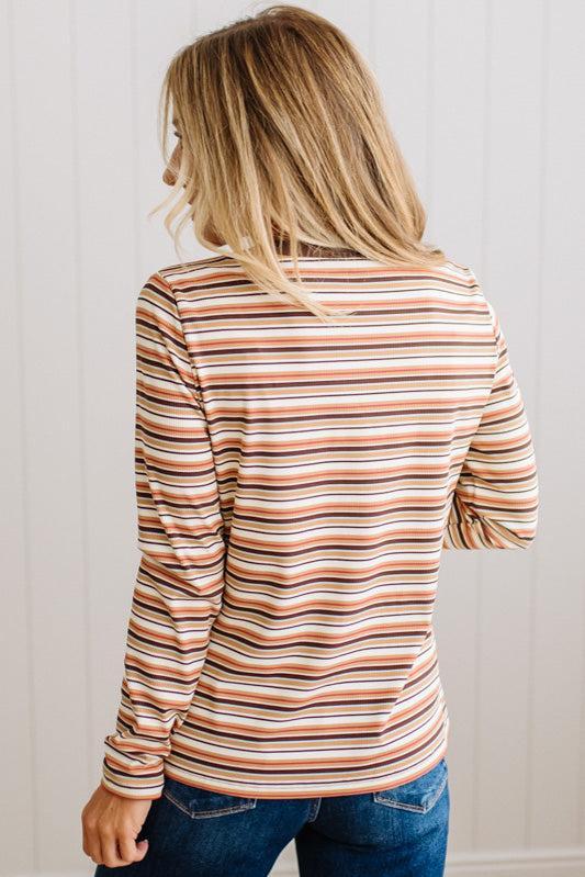 A woman viewed from behind wearing a long-sleeve striped top with horizontal stripes in terracotta orange, brown, beige, and white, paired with blue jeans against a white paneled wall.