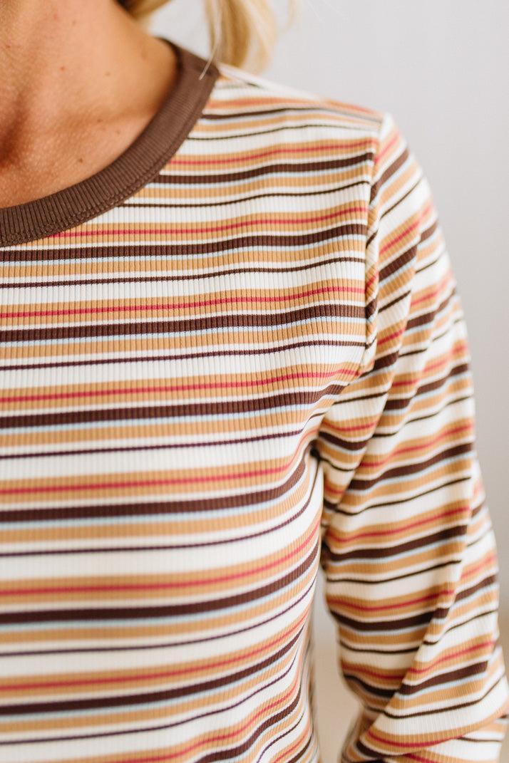 Close-up of a person wearing a long-sleeve ribbed top with horizontal stripes in brown, beige, orange, and white, featuring a solid brown crew neck.