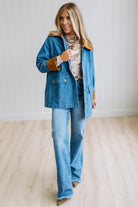 A woman modeling a blue denim jacket with brown suede-like trim over a graphic tee and blue jeans, standing in a bright room with light wood floors and white paneled walls.