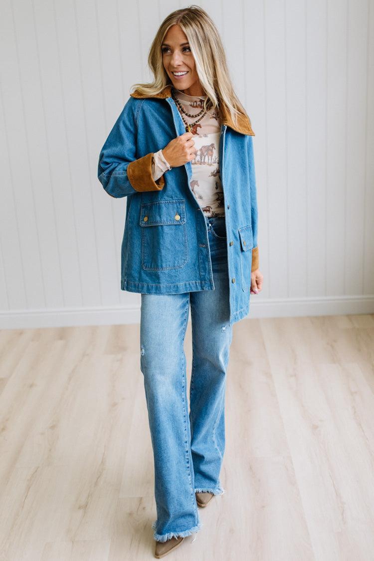 A woman modeling a blue denim jacket with brown suede-like trim over a graphic tee and blue jeans, standing in a bright room with light wood floors and white paneled walls.
