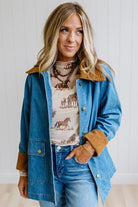 A woman wearing a blue denim jacket with brown suede-like collar and cuffs, layered over a western-print top and jeans, standing against a white paneled wall.