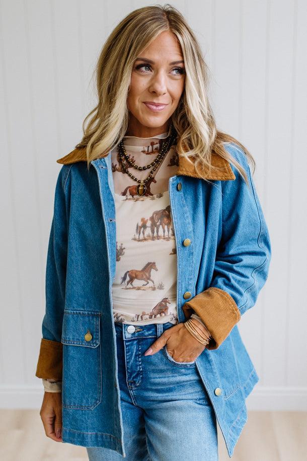 A woman wearing a blue denim jacket with brown suede-like collar and cuffs, layered over a western-print top and jeans, standing against a white paneled wall.