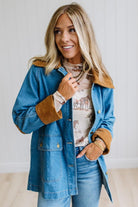 A woman wearing a blue denim jacket with tan suede-like collar and cuffs, layered over a graphic t-shirt and blue jeans, standing against a white paneled wall.
