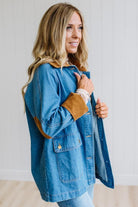 A woman wearing a blue denim jacket with brown suede-like trim on the collar and cuffs, standing in a bright indoor setting with a white paneled wall.