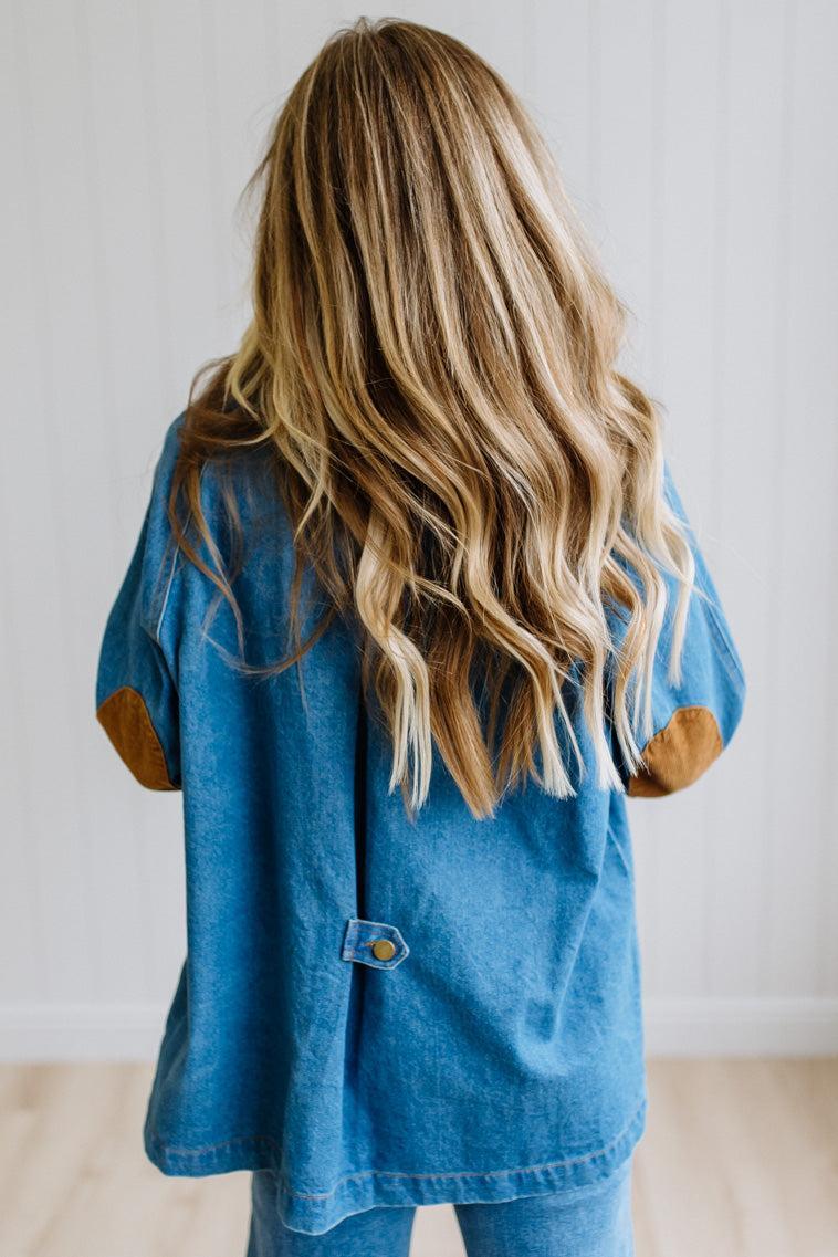 A child viewed from behind wearing a blue denim shirt with brown elbow patches, standing against a white paneled wall.