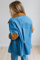 A woman shown from the back wearing a blue denim jacket with tan corduroy collar and elbow patches, paired with blue jeans, against a white paneled wall and light wood floor.