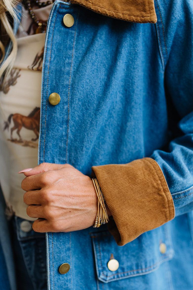 Close-up of a person wearing a blue denim jacket with a brown corduroy collar and pocket, paired with a patterned cream top and gold bracelets.