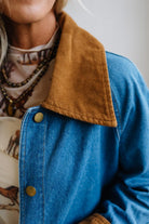 Close-up of a person wearing a blue denim jacket with a wide brown corduroy collar and brass-tone snap buttons, layered over a patterned top.