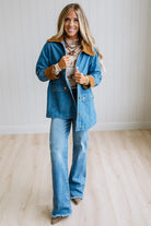 A woman modeling a blue denim jacket with a brown suede collar and cuffs, paired with matching blue denim flare jeans, standing in a bright room with light wood floors and white paneled walls.