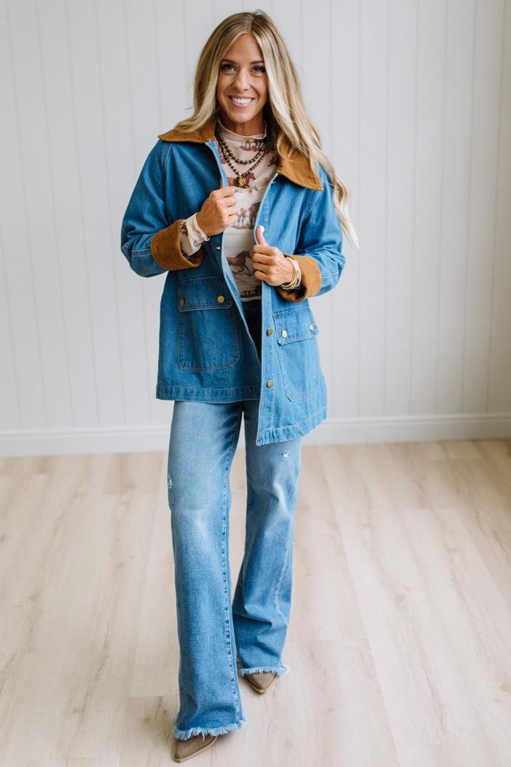 A woman modeling a blue denim jacket with a brown suede collar and cuffs, paired with matching blue denim flare jeans, standing in a bright room with light wood floors and white paneled walls.