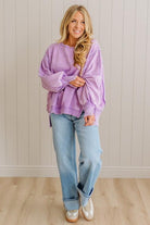 Woman wearing a purple oversized sweater and blue jeans standing against a light wooden paneled wall.