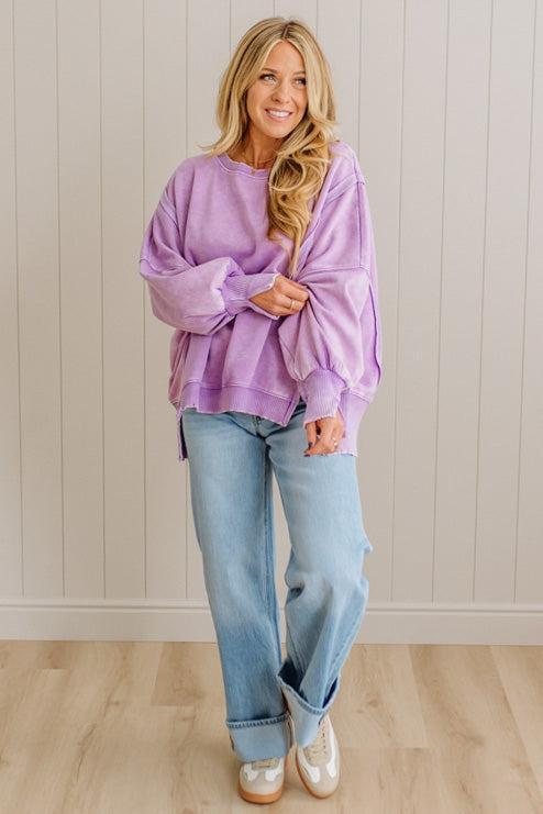 Woman wearing a purple oversized sweater and blue jeans standing against a light wooden paneled wall.