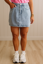 Person wearing a denim skirt and white sneakers in a room with wooden flooring and a neutral wall.