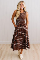 A woman modeling a sleeveless brown midi dress with white polka dots, featuring a smocked bodice and side pockets, paired with cream ankle boots against a white paneled wall and light wood floor.